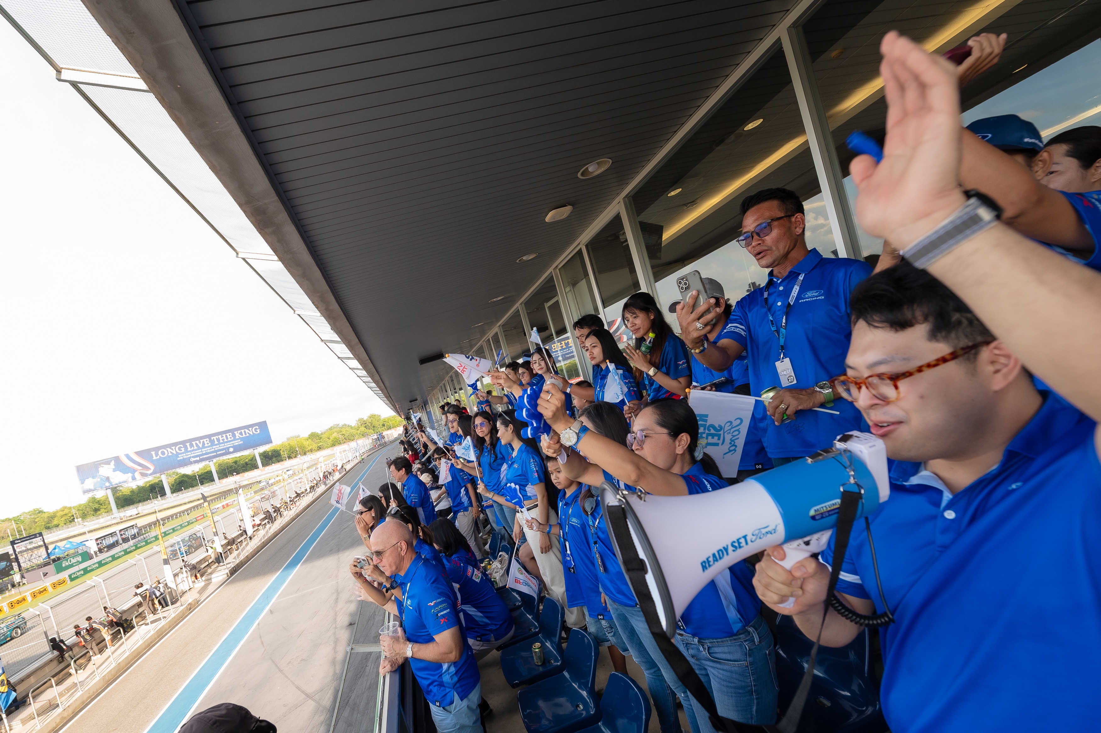 Enthusiastic fans in blue Ford shirts, waving Ready Set Ford flags and cheering as a race truck speeds past.
